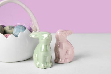 Easter bunnies and basket with painted eggs on table against lilac background