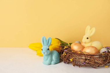 Easter bunnies, tulip flowers and nest with painted eggs on table against yellow background