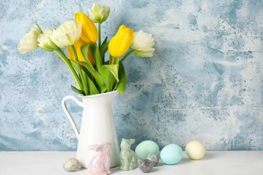 Easter bunnies, vase with tulip flowers and painted eggs on table near blue grunge wall