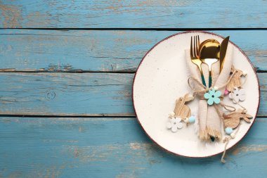 Table setting with Easter bunnies and cutlery in napkin on blue wooden background