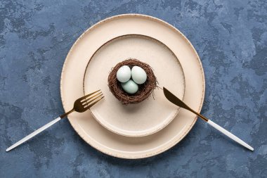 Table setting with cutlery and Easter eggs in nest on blue grunge background