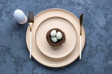 Table setting with cutlery and Easter eggs in nest on blue grunge background