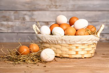 Wicker basket with Easter eggs and hay on wooden table near grey wall