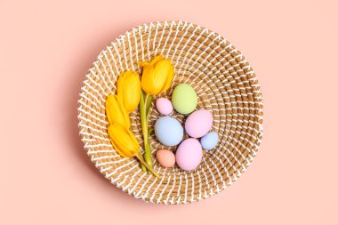 Wicker basket with painted Easter eggs and tulip flowers on pink background