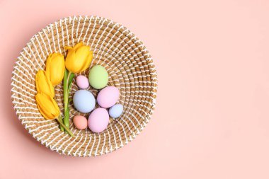 Wicker basket with painted Easter eggs and tulip flowers on pink background