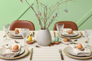 Festive dining table with eggs and pussy willow branches served for Easter celebration near green wall