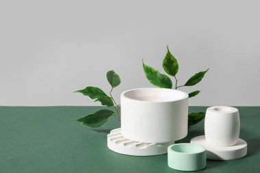 Plaster podiums and ficus branches on green table against grey background