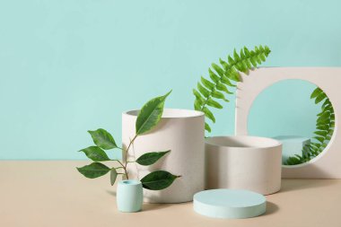 Plaster podiums with ficus and fern branches on beige table against turquoise background
