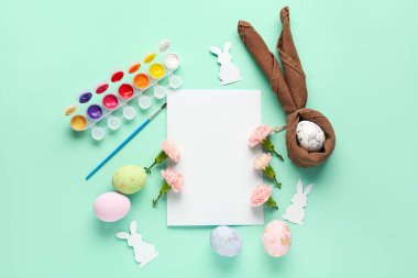 Blank paper sheet with flowers, paints and Easter eggs on green background