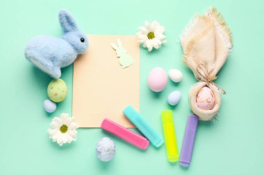 Blank paper sheet with rabbits, Easter eggs, flowers and markers on green background