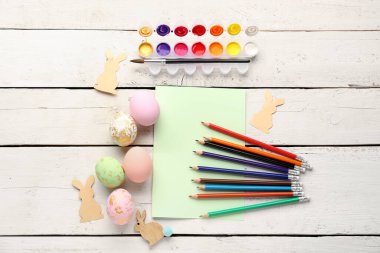 Blank card with pencils, Easter eggs, rabbits and paints on white wooden background