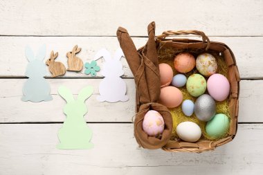Basket with Easter eggs and paper rabbits on white wooden background