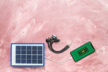 Portable solar panel charging mobile phone and different cables on pink grunge background