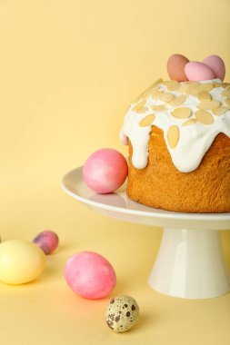 Stand with Easter cake and painted eggs on yellow background
