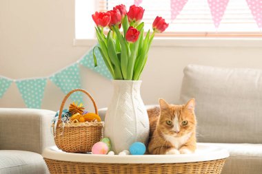 Cute cat, vase with tulips and Easter eggs on table at home