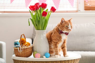 Cute cat, vase with tulips and Easter eggs on table at home