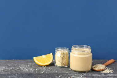 Jars with tasty tahini, sesame seeds and lemon slice on dark wooden table near color wall