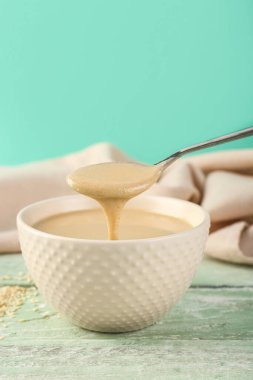 Pouring of tasty tahini from spoon into bowl on table near color wall