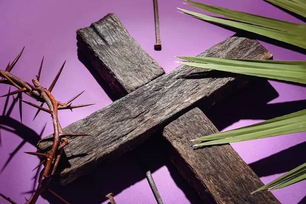 Wooden cross, crown of thorns, nails and palm leaf on purple background, closeup. Good Friday concept