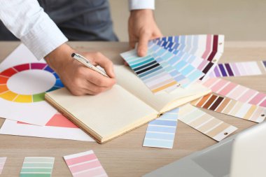 Male painter with color palettes writing in notebook at table, closeup