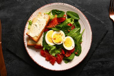 Plate of delicious salad with boiled eggs, salmon and cream cheese sandwiches on black grunge background