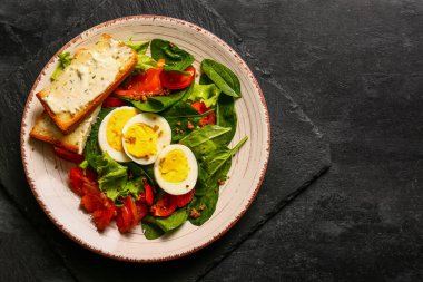 Plate of delicious salad with boiled eggs, salmon and cream cheese sandwiches on black grunge background