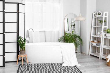 Interior of light bathroom with bathtub, shelving units and houseplants
