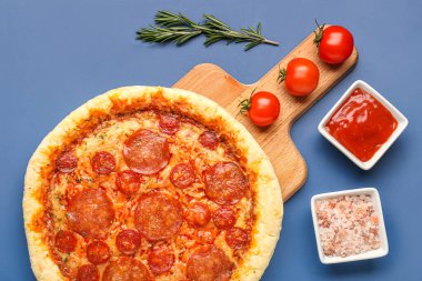 Board with delicious pepperoni pizza and ingredients on blue background