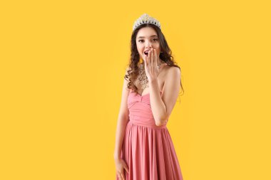 Beautiful surprised young woman in stylish prom dress and tiara on yellow background