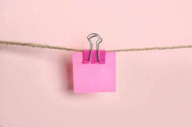 Sticky note with binder clip hanging on rope against pink background, closeup