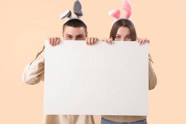 Happy young couple in bunny ears with blank poster on beige background. Easter celebration