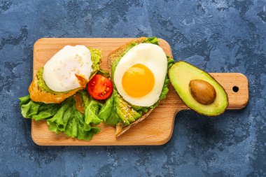 Board with delicious avocado toasts and eggs on blue grunge background