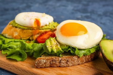 Board with delicious avocado toasts and eggs on blue grunge background, closeup