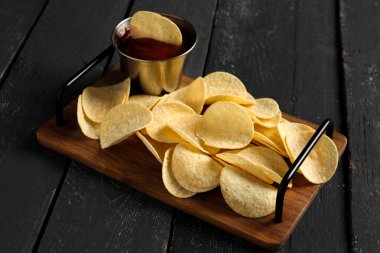 Board with delicious potato chips and bowl of ketchup on black wooden background