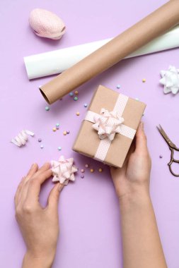 Woman with gift box, bows and wrapping paper on lilac background