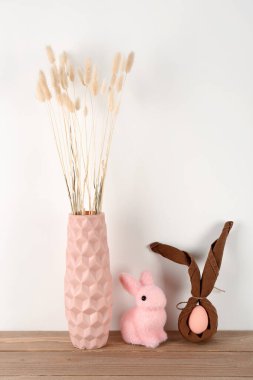 Vase with dried flowers, Easter egg and rabbit on table near white wall