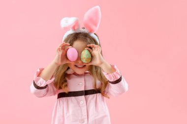 Cute little girl in bunny ears with painted Easter eggs on pink background