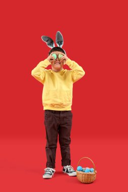Cute little boy with bunny ears and Easter basket on red background