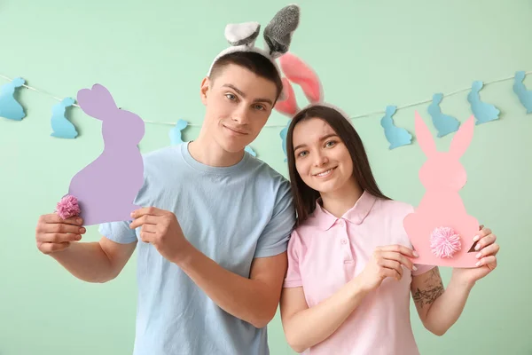Happy young couple in bunny ears with paper Easter rabbits near green wall