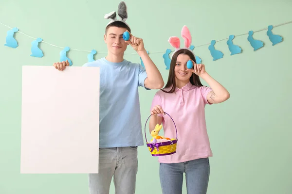 Happy young couple in bunny ears with Easter eggs and blank poster near green wall