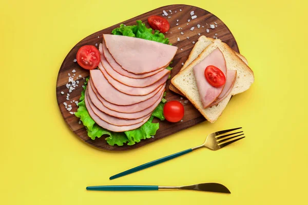 Board with slices of tasty ham, bread pieces, lettuce and tomatoes on yellow background