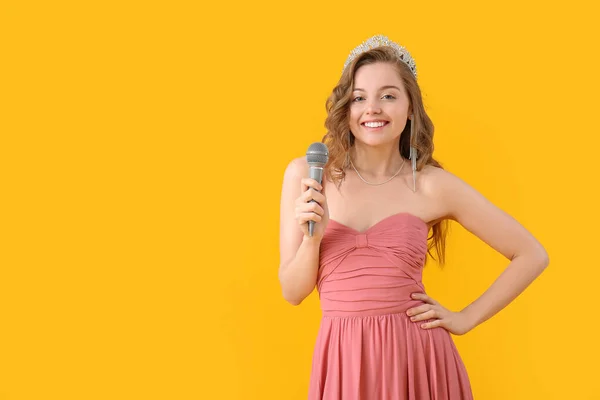 Young woman in tiara and prom dress with microphone on yellow background