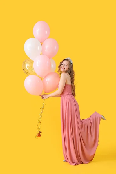 Young woman in prom dress with balloons on yellow background