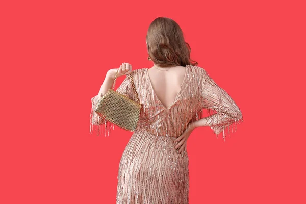 Young woman in shiny prom dress with bag on red background, back view