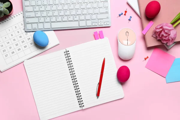 Blank notebook with office stationery, computer keyboard, flower and Easter eggs on pink background