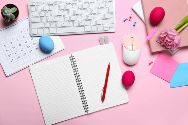 Blank notebook with office stationery, computer keyboard, flower and Easter eggs on pink background