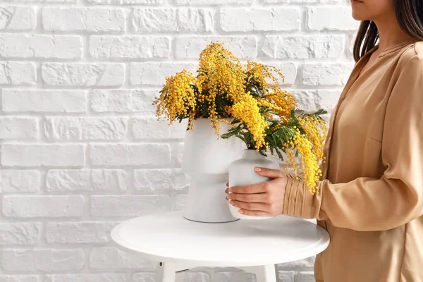 Woman holding vase with mimosa flowers on table near light brick wall in room
