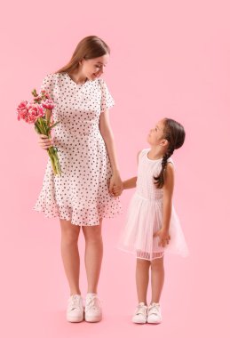 Sevimli küçük kız ve çiçekli annesi pembe arka planda el ele tutuşuyorlar.