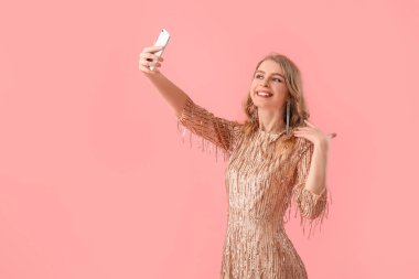 Teenage girl in shiny prom dress taking selfie on pink background
