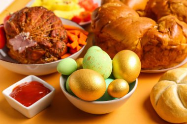 Bowl of painted eggs and tasty dishes for Easter dinner on color background, closeup